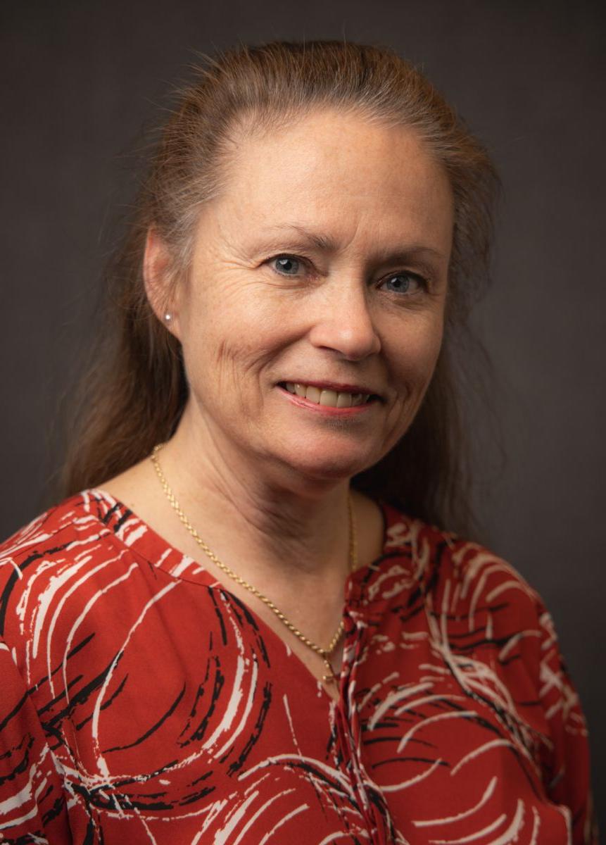 A Caucasian female with long brown hair, Gail Volz smiles at the camera and is wearing a red top with a black and white paint circle pattern on it as well as a chain necklace.